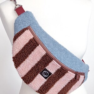 May include: A crescent-shaped fanny pack made of textured sherpa fabric. The bag features a light blue top panel and a front section with alternating vertical stripes of light pink and dark brown. A small black square patch with a white smiley face icon is centered on the front. It includes a dark red adjustable strap with a silver metal clip.