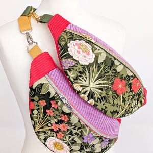 May include: A floral print fanny pack with a red corduroy accent and a purple corduroy center. The bag features a zipper closure and an adjustable strap with gold-colored hardware. The floral pattern includes pink, white, and purple flowers.