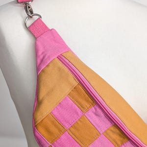 Könnte beinhalten: Eine pinkfarbene und beige Bauchtasche mit Schachbrettmuster. Die Tasche hat einen pinkfarbenen Reißverschluss und Gurt und wird auf einer Schaufensterpuppe gezeigt. Die Tasche ist aus Cordstoff.