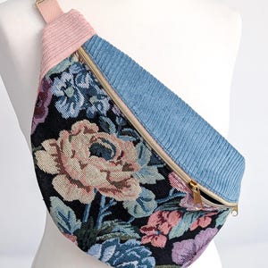 May include: A floral print fanny pack with a light blue corduroy top panel and a pink corduroy strap. The bag features a gold zipper and hardware. The floral pattern includes pink, blue, and purple flowers on a black background.