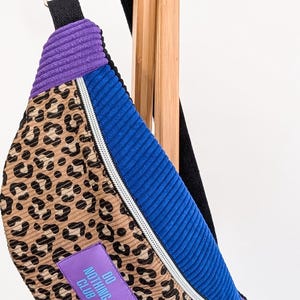 Könnte beinhalten: Eine Bauchtasche aus Cord mit Leopardenmuster, blauen und lila Akzenten. Die Tasche hat einen silbernen Reißverschluss und ein kleines lila Etikett mit der Aufschrift "DO NOTHING CLUB". Die Tasche hängt an einem Holzgestell.