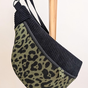 May include: A black and olive green fanny pack with a leopard print design. The top section is black corduroy, and the bag has a black strap and a green zipper. The bag is hanging on a wooden rack.