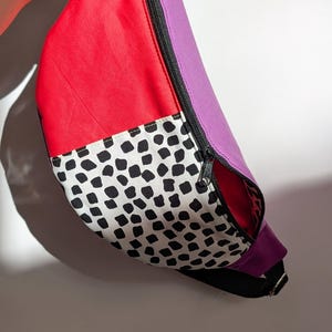 May include: A colorful fanny pack with a black zipper. The bag features a red panel, a purple panel, and a white panel with black spots. The bag is held by a silver key ring.