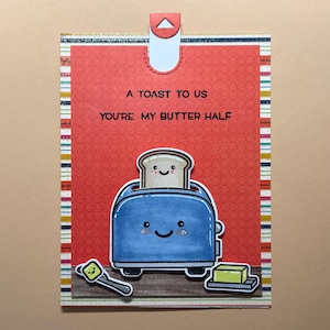 May include: A greeting card with a red background and the text "A toast to us, you're my butter half." The card features a cartoon toaster with a slice of toast and a stick of butter. The card has a pull tab.