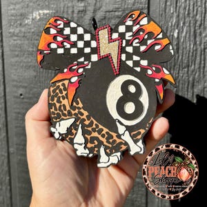 May include: A decorative ornament shaped like a bow featuring a black eight ball with a white number eight, a leopard print section, white skeleton hand accents, a black and white checkered pattern, orange flames, and a gold glitter lightning bolt.
