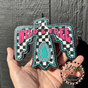 May include: A thunderbird-shaped hanging ornament featuring a black and white checkered pattern, bright pink text reading WILD & FREE, and a central turquoise glitter teardrop accent. The edges are lined with small, raised turquoise dots.