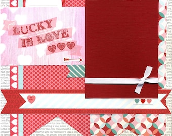 May include: A scrapbook page with a red, white, and pink color scheme. The page features a large red frame with a white ribbon and bow. The text "LUCKY IN LOVE" is written in red and white letters on a pink background. The page also includes various hearts, arrows, and other decorative elements.