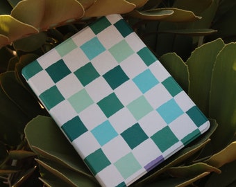 May include: A white notebook with a checkered pattern in shades of green and blue. The notebook is lying on top of green leaves.