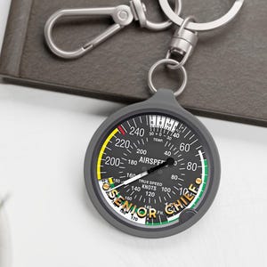 May include: A circular keychain featuring an aviation airspeed indicator gauge design with a black face, white markings, and a needle. The gauge includes the text AIRSPEED, TRUE SPEED KNOTS, and SENIOR CHIEF in yellow lettering at the bottom.
