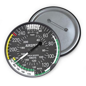 May include: A round, black and silver button with an airspeed gauge design. The gauge displays numbers and markings in white, green, and yellow, with the words "AIRSPEED" and "TRUE SPEED KNOTS" visible. The button has a pin on the back.