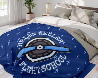 Puede incluir: Una manta de forro polar azul oscuro con un patrón repetitivo de pequeños aviones azul claro. En el centro, un logotipo circular muestra una hélice de avión estilizada sobre una rueda gris oscuro, con el texto HELEN KELLER FLIGHT SCHOOL.