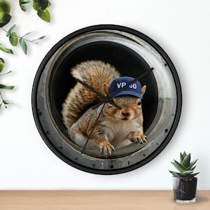 May include: Round wall clock with a black frame featuring a squirrel wearing a blue cap with "VP 00" printed on it. The squirrel is inside a circular opening. The clock hands are black.