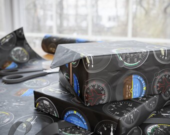 May include: Wrapping paper featuring a repeating pattern of black and white aviation instrument gauges, including altimeters and artificial horizons with blue and brown accents. The paper is folded over a rectangular box, displaying detailed dials.