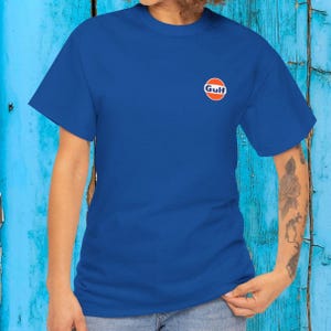 Pode incluir: Uma t-shirt de manga curta azul-real com um pequeno logótipo circular da Gulf na parte superior esquerda do peito. O logótipo consiste num círculo branco com rebordo azul, contendo a palavra Gulf em azul sobre uma faixa horizontal laranja.