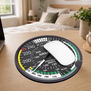 May include: A round mouse pad featuring a black and white aviation airspeed indicator gauge graphic with yellow and green arcs, white numerical markings, and the text AIRSPEED, TRUE SPEED, KNOTS, and M.P.H. A white computer mouse rests on top.