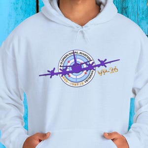 Puede incluir: Una sudadera con capucha blanca que presenta un gráfico de un avión P-3 Orion morado sobre un objetivo de radar circular. El texto dice P-3 ORION NAVAL AVIATION SUB-HUNTER VETERAN y VP-26 en letras doradas. El diseño está centrado en el pecho.