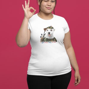 May include: White t-shirt featuring a dog dressed as an archer with the text "I get that dog in me." The dog wears a green and brown hat and holds arrows. The person is winking and making an "okay" hand gesture.
