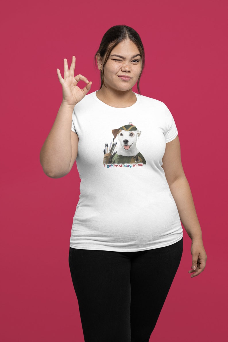 May include: White t-shirt featuring a dog dressed as an archer with the text "I get that dog in me." The dog wears a green and brown hat and holds arrows. The person is winking and making an "okay" hand gesture.