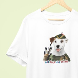 May include: White t-shirt featuring a Jack Russell Terrier dressed as Robin Hood with a quiver of arrows. The text "I got that dog in me" is printed below the dog in colorful letters. The shirt is hanging on a wooden hanger.