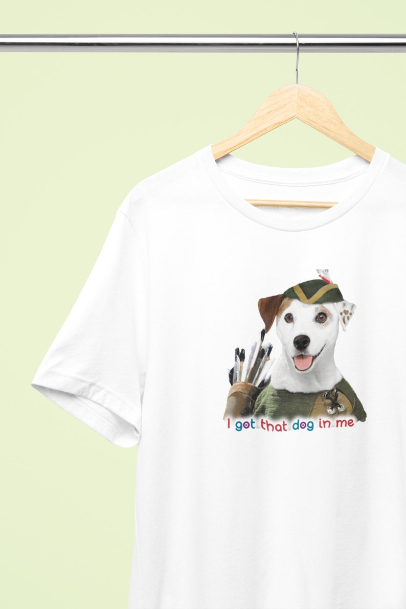 May include: White t-shirt featuring a Jack Russell Terrier dressed as Robin Hood with a quiver of arrows. The text "I got that dog in me" is printed below the dog in colorful letters. The shirt is hanging on a wooden hanger.