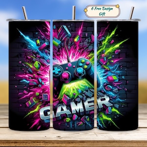Puede incluir: Un vaso negro con un diseño vibrante que presenta un mando de videojuegos y la palabra "GAMER" en negrita. El diseño incorpora colores neón rosa, azul y verde, con un fondo de pared de ladrillos. Incluye una pajita y una etiqueta de regalo.