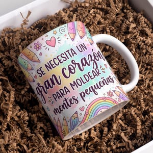 Puede incluir: Taza de cerámica blanca con un diseño colorido con lápices, corazones y un arcoíris. La taza tiene el texto "SE NECESITA UN gran corazón PARA MOLDEAR mentes pequeñas". La taza está dentro de una caja blanca con papel marrón.
