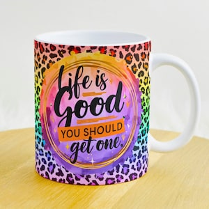 May include: A white ceramic mug with a rainbow-colored background and a leopard print pattern. The mug has the text "Life is good you should get one." in black lettering.