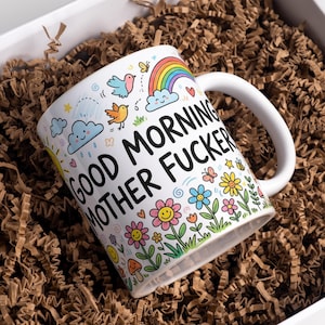 May include: White ceramic mug with the text "GOOD MORNING MOTHER FUCKER" in black. The mug features a colourful design of a rainbow, birds, clouds, flowers, and butterflies. The mug is inside a white box with brown paper filler.
