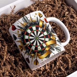 May include: White ceramic mug featuring a dartboard design with darts, gold ribbons, and stars. The mug has a white handle and is set in a white box with brown paper filler. The mug is a gift for a darts player.