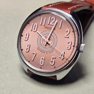 Peut inclure: Une montre-bracelet avec un boîtier rond argenté et un cadran cuivré doté de chiffres arabes blancs. Le cadran affiche le texte "PAKETA" et "СДЕЛАНО В СССР" avec un emblème d'étoile centrale. Elle comprend un bracelet en cuir marron avec surpiqûres.