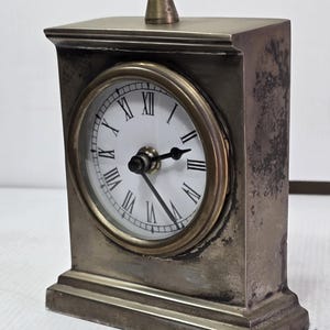 Può includere: Un orologio da tavolo rettangolare in metallo color ottone con finitura usurata. L'orologio presenta un quadrante bianco circolare con numeri romani neri, due lancette nere e un piccolo anello di presa sulla parte superiore.