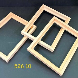 May include: Three unfinished rectangular wooden frames are arranged on a dark surface. The frames are made of light-colored wood and are empty. The number "526 10" is printed in yellow on the surface.