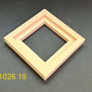 May include: A square, light-colored wooden frame with mitered corners and a stepped inner edge. The frame is positioned at an angle, and the yellow text 1026 19 is visible in the lower left corner of the image.