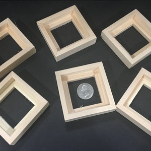 May include: Six unfinished, square wooden frames are arranged on a dark surface. One frame contains a quarter coin. The frames are made of light-colored wood and have a recessed square opening in the center. The frames are likely for small artwork or crafts.