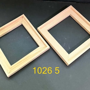May include: Two square, light-colored wooden frames with a natural grain finish and angled inner edges. The frames are positioned diagonally, and the yellow text 1026 5 is displayed between them. The frames have a simple, clean construction.