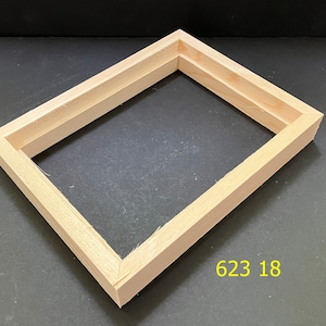 May include: A rectangular, unfinished light-colored wooden frame with mitered corners and a recessed inner edge. The frame features a visible wood grain texture and is positioned at an angle. The yellow text 623 18 is printed near the bottom right.