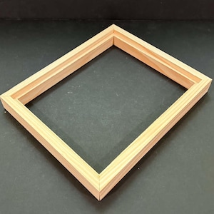 May include: A rectangular picture frame made of light-colored, unfinished natural wood. The frame features a simple, clean-lined profile with mitered corners, showcasing the visible grain and texture of the wood material in a light tan hue.