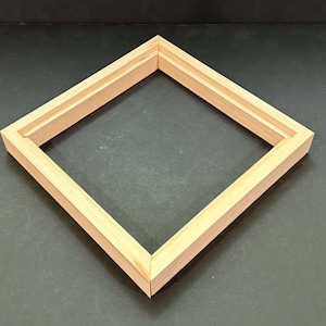 May include: A square wooden floating frame with a natural light brown finish. The frame features a clean, simple design with mitered corners and a recessed inner edge, providing a structured border for mounting artwork or canvas panels.