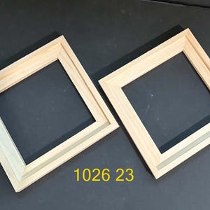 May include: Two square, unfinished light-colored wooden frames with mitered corners and beveled inner edges. The frames are positioned diagonally, and the yellow text 1026 23 is printed in the lower center area between the two wooden frames.