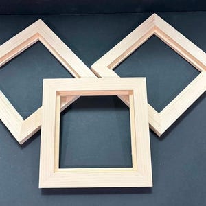 May include: Three square, light-colored wooden frames arranged in a triangular formation. Each frame features a smooth, natural wood grain finish with a recessed inner edge designed for mounting artwork or canvases within the frame structure.