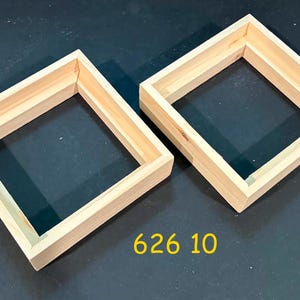 May include: Two square, light-colored wooden frames with mitered corners and a recessed inner edge. The frames are constructed from natural, unfinished wood. The text 626 10 is printed in yellow below the frames on the dark surface.