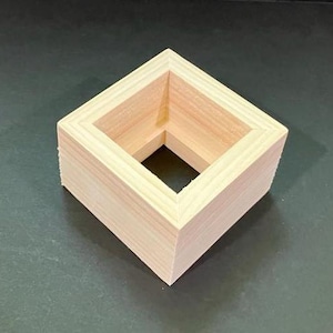 May include: A small, square wooden box with an open top and bottom. The box is made of light-colored wood and has a simple, geometric design. The box is sitting on a dark surface.