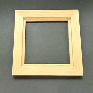 May include: A square, light-colored wooden picture frame with a smooth, natural finish. The frame features a wide, flat border with beveled inner edges that slope inward toward the center opening, creating a simple and clean geometric design.