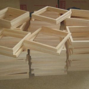 May include: A large collection of square, unfinished light-colored wooden frames stacked in multiple piles. Each frame features a simple, clean-edged construction with visible wood grain, designed for mounting canvas or other display materials.