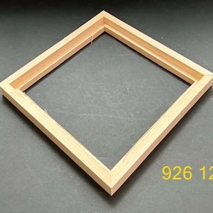 May include: A square, light-colored wooden frame with a mitered corner design and a recessed inner edge. The frame is positioned at an angle, and the yellow text 926 12 is visible in the lower right corner of the image area.