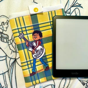 May include: A black Kindle e-reader next to a yellow, green, and white plaid e-reader case. The case features a cartoon image of a boy playing a guitar. The case has a white flap with a yellow button closure.