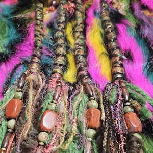 Puede incluir: Primer plano de tres extensiones de rastas con cuentas de madera y detalles de piedra marrón rojizo. Las rastas son marrones y verdes, con un fondo colorido de piel sintética rosa, amarilla, verde y negra.