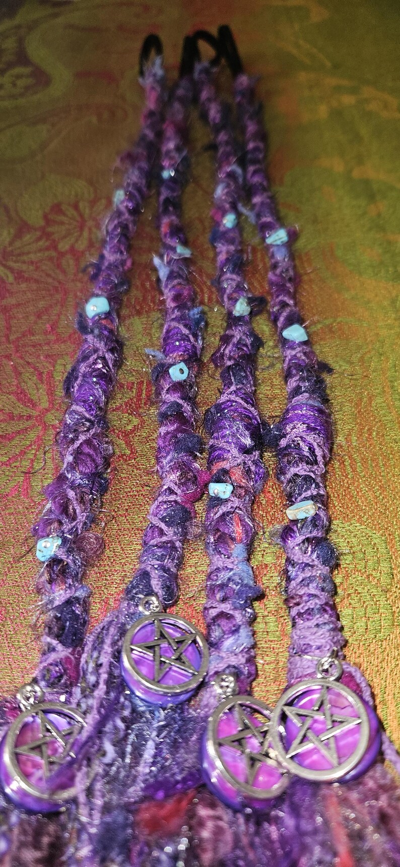 Puede incluir: Extensiones de rastas moradas y azules con cuentas turquesas y dijes de pent&aacute;culo plateado. Las rastas est&aacute;n hechas de un material texturizado y tejido. Los dijes son redondos con un centro morado y un dise&ntilde;o de pent&aacute;culo plateado.