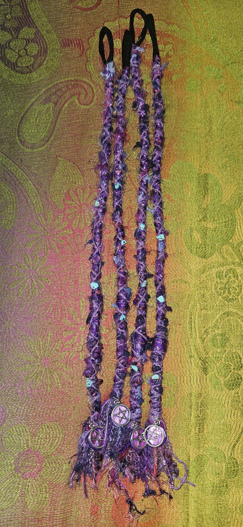 Puede incluir: Extensiones de rastas moradas y violetas con cuentas turquesas y colgantes de pentagrama plateados. Las rastas est&aacute;n unidas a bandas el&aacute;sticas negras. El fondo es un estampado de cachemira en tonos dorados, verdes y rosas.