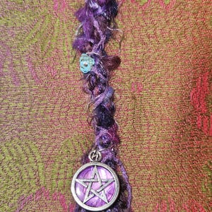 Puede incluir: Una envoltura de rastas morada hecha a mano con un dije de pentagrama plateado. La envoltura est&aacute; hecha de varios tonos de hilo morado y presenta una peque&ntilde;a cuenta turquesa. El fondo es una tela estampada en tonos verdes y rosas.